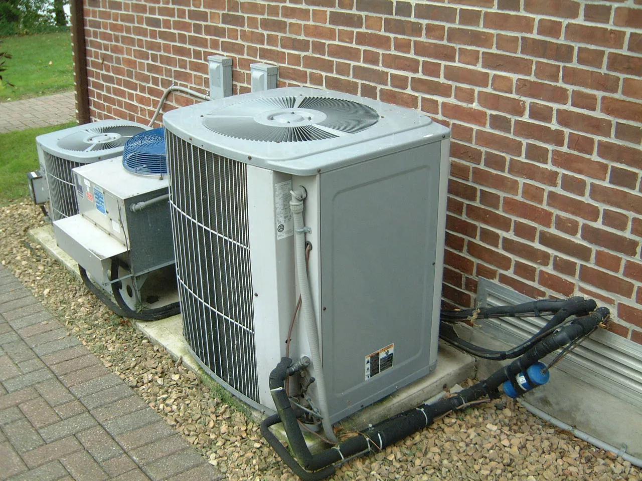 Two outdoor air conditioner condensers on a pad beside a red brick house wall with insulated refrigerant lines