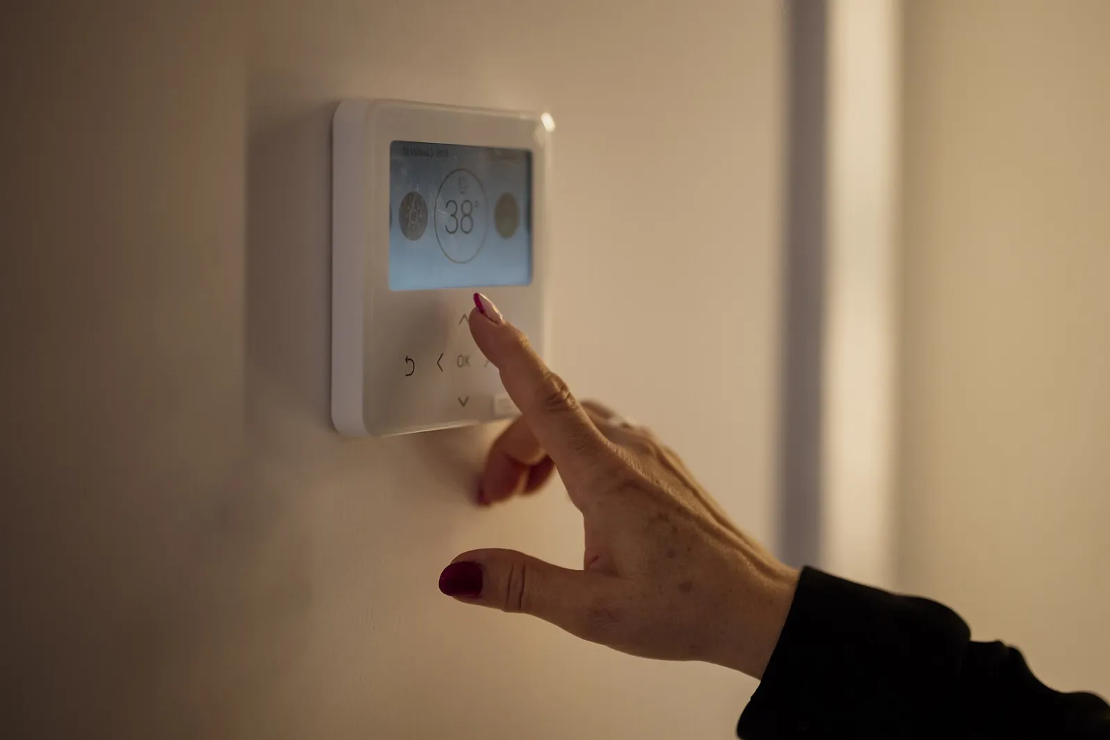 Hand pressing the buttons on a white digital wall thermostat in a dim hallway